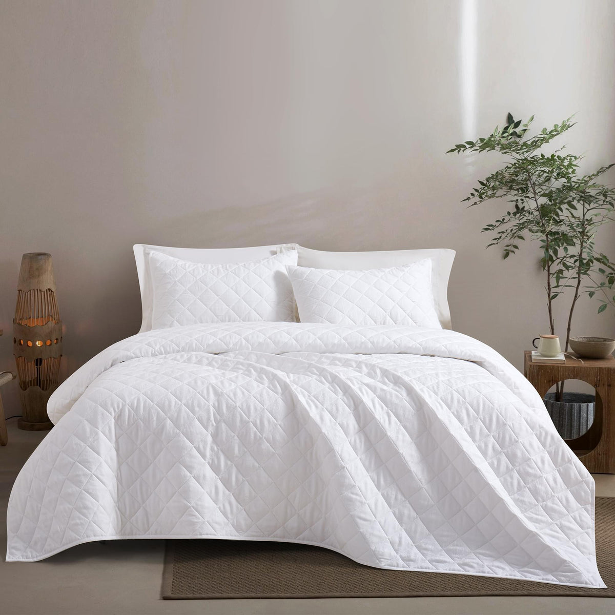 White Timeless Heritage Woven Quilt Set White comforter on a bed in a bedroom setting with a plant and side table.