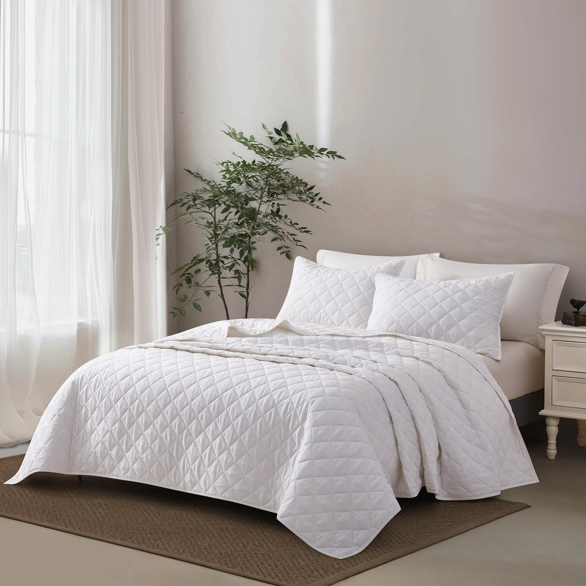 White Timeless Heritage Woven Quilt Set White on a bed in a bedroom with a plant and nightstand.