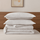 Stack of light gray pillows and duvet on a bed with a wooden headboard and abstract art in the background.