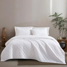 White Timeless Heritage Woven Quilt Set White comforter on a bed in a bedroom setting with a plant and side table.