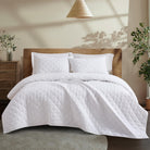 White Aurora Bloom Leaf Jacquard Quilt Set White on a bed with wooden headboard in a room with plants and a lamp.