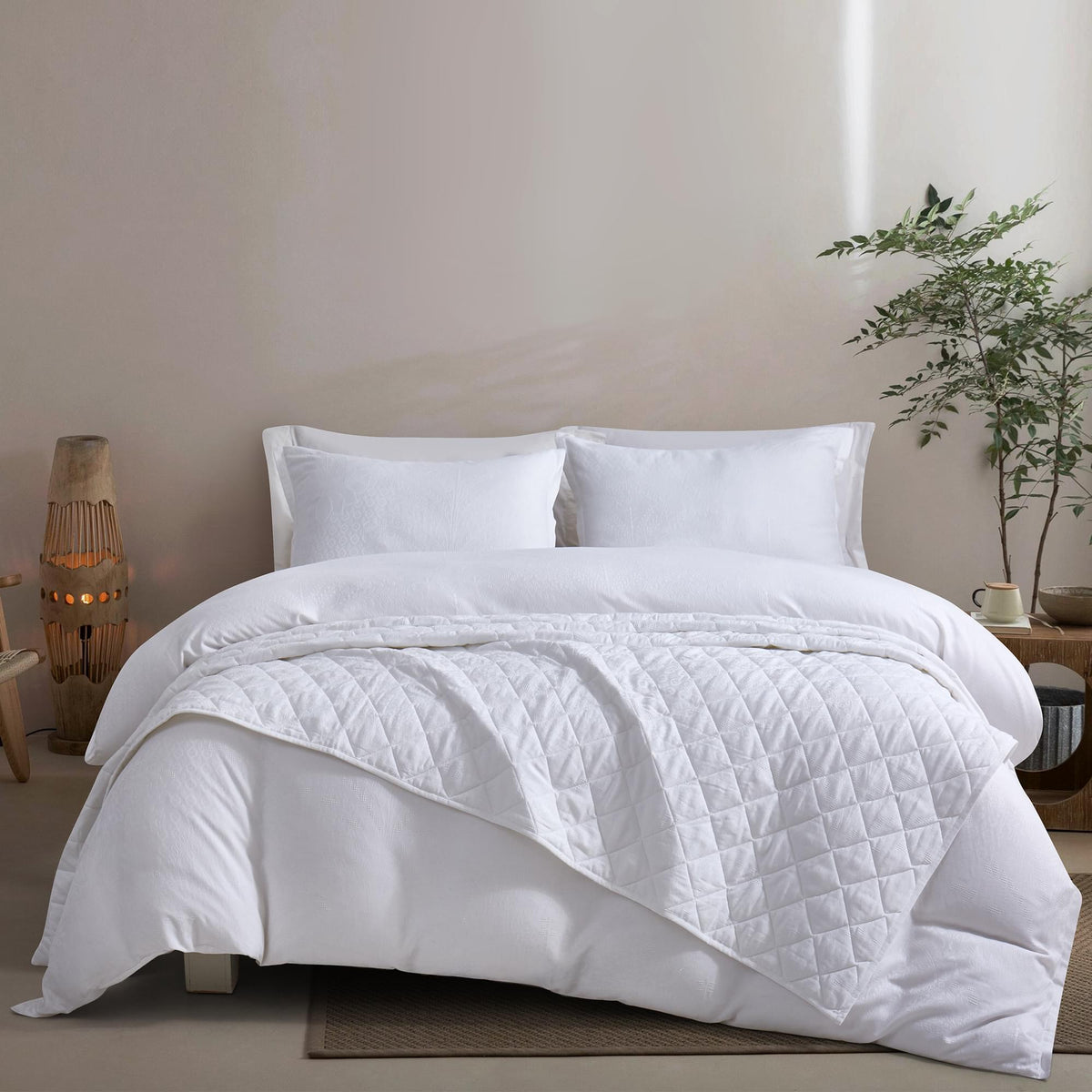 White Timeless Heritage Woven Quilt Set White comforter on a bed in a bedroom setting with a plant and lamp.
