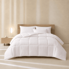 A neatly made bed with a white All Season Bamboo Duvet White Insert and two white pillows, set against a light beige wall with a wooden headboard. A small bedside table with a lit lamp sits on the left.