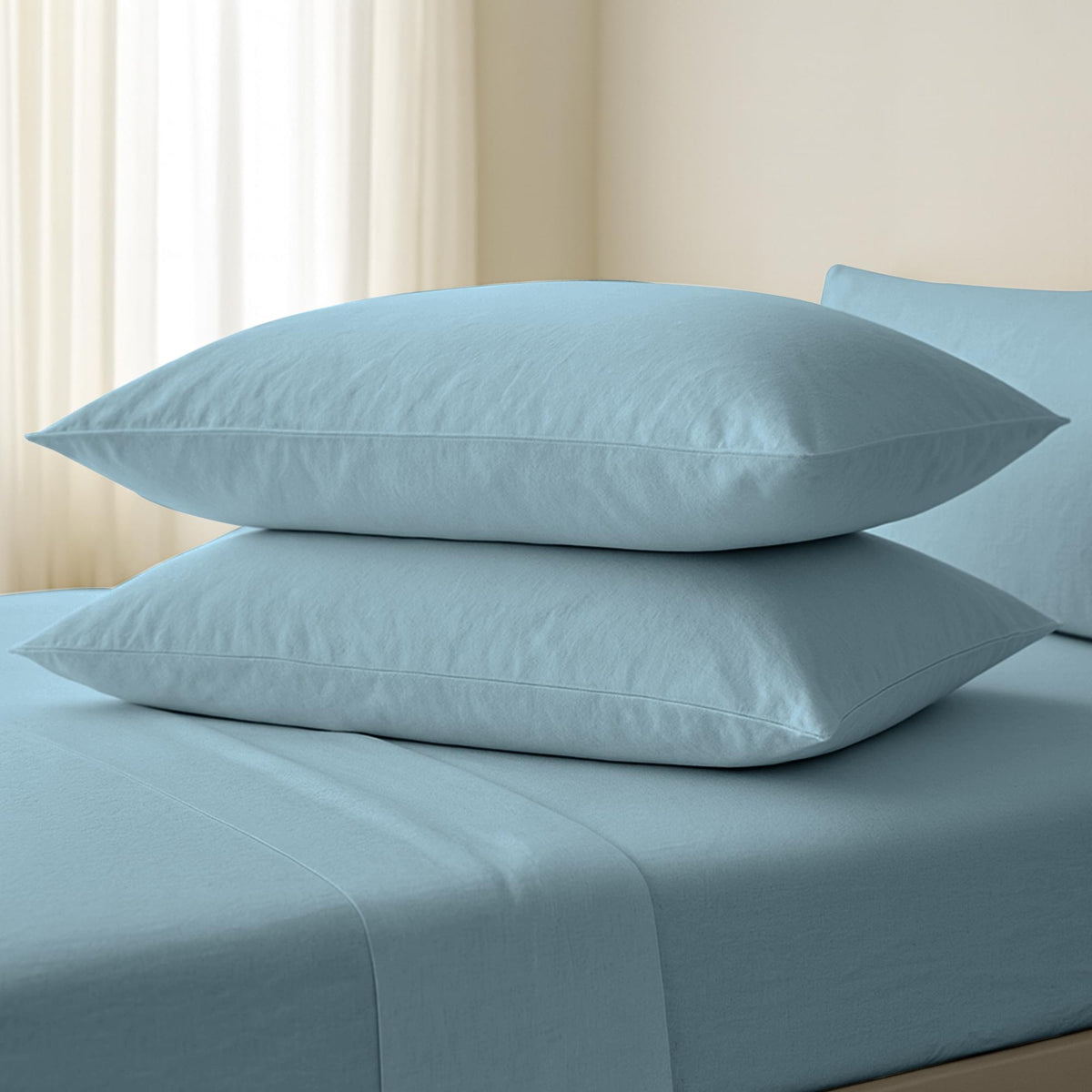 Two light blue pillows on a matching bed sheet with a neutral curtain background