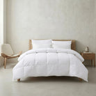 A white quilted Down and Feather Duvet with matching white pillows on a light wood bed frame, styled in a minimalist bedroom with a beige textured wall, a wooden chair, and a small wooden nightstand.