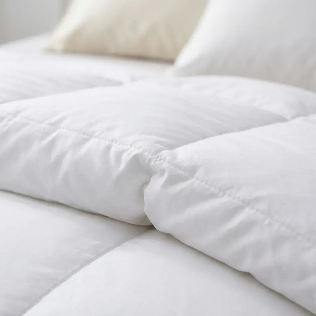 A close-up of a white quilted Down and Feather Products Duvet on a bed, with soft white pillows visible in the background.