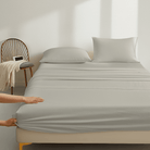 Person adjusting gray fitted sheet on a bed in a minimalistic room.