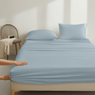 Person adjusting light blue bedding on a bed in a room with a chair and window.