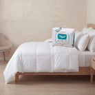 A White Goose Feather Duvet set on a wooden bed frame with matching white pillows, alongside its white packaging bag featuring a blue whale graphic.