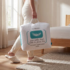 A person in cream knit pajamas carries a large white Newseason goose feather duvet packaging bag with teal accents in a bright bedroom with wooden floors and a bed.