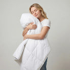 A woman hugging a large white quilted comforter or duvet against a plain light gray studio background.