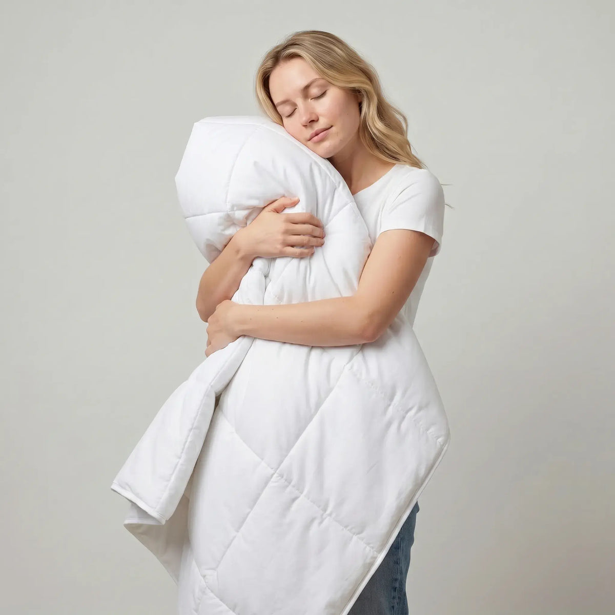 A woman hugging a large white quilted comforter or duvet against a plain light gray studio background.