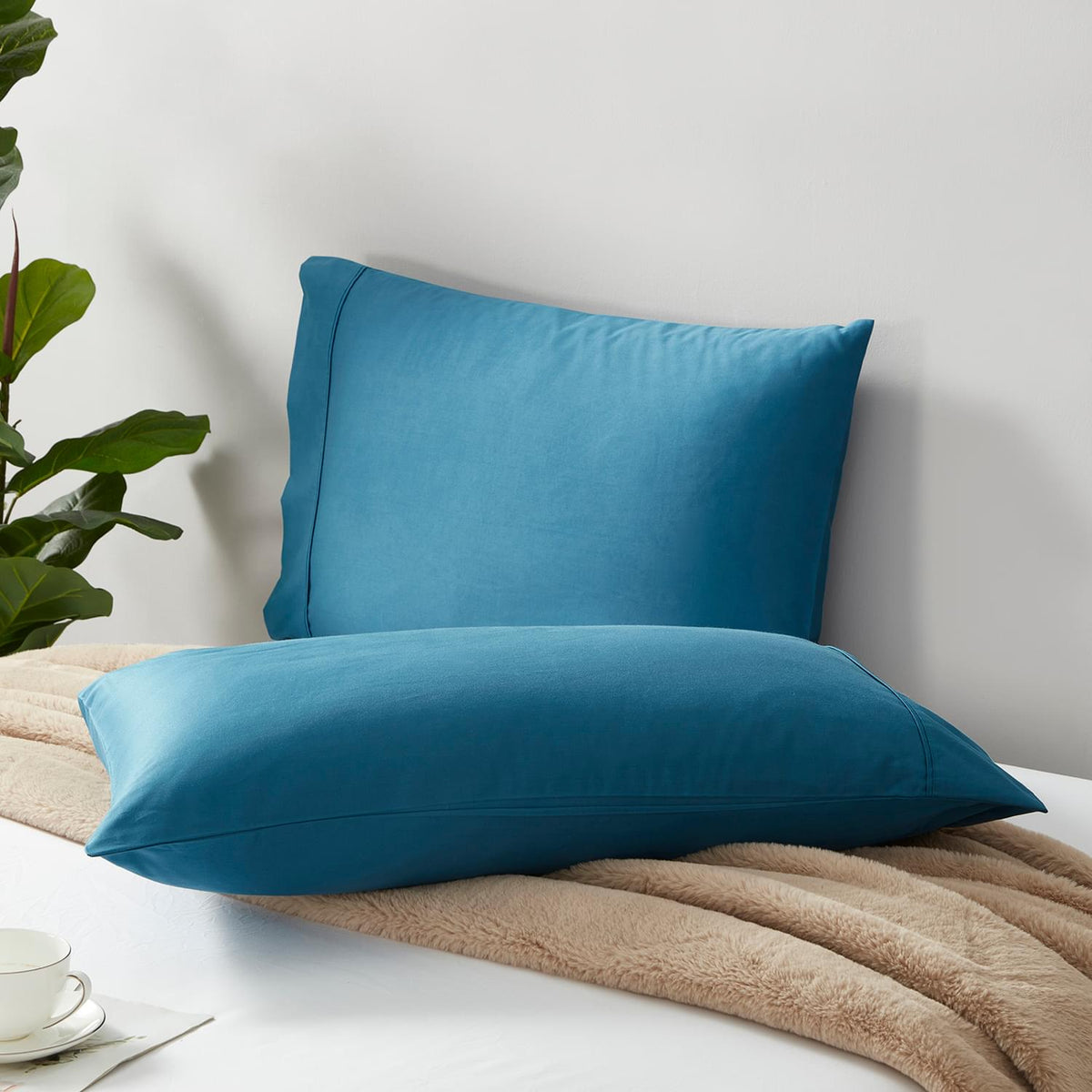 Two blue Classic 500 Thread Count Organic Pillowcase Pair on a bed with a plant and cup in the background