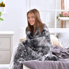 Woman sitting on a bed wrapped in a Hug Me Faux Weighted Throw Blanket in a bright room.